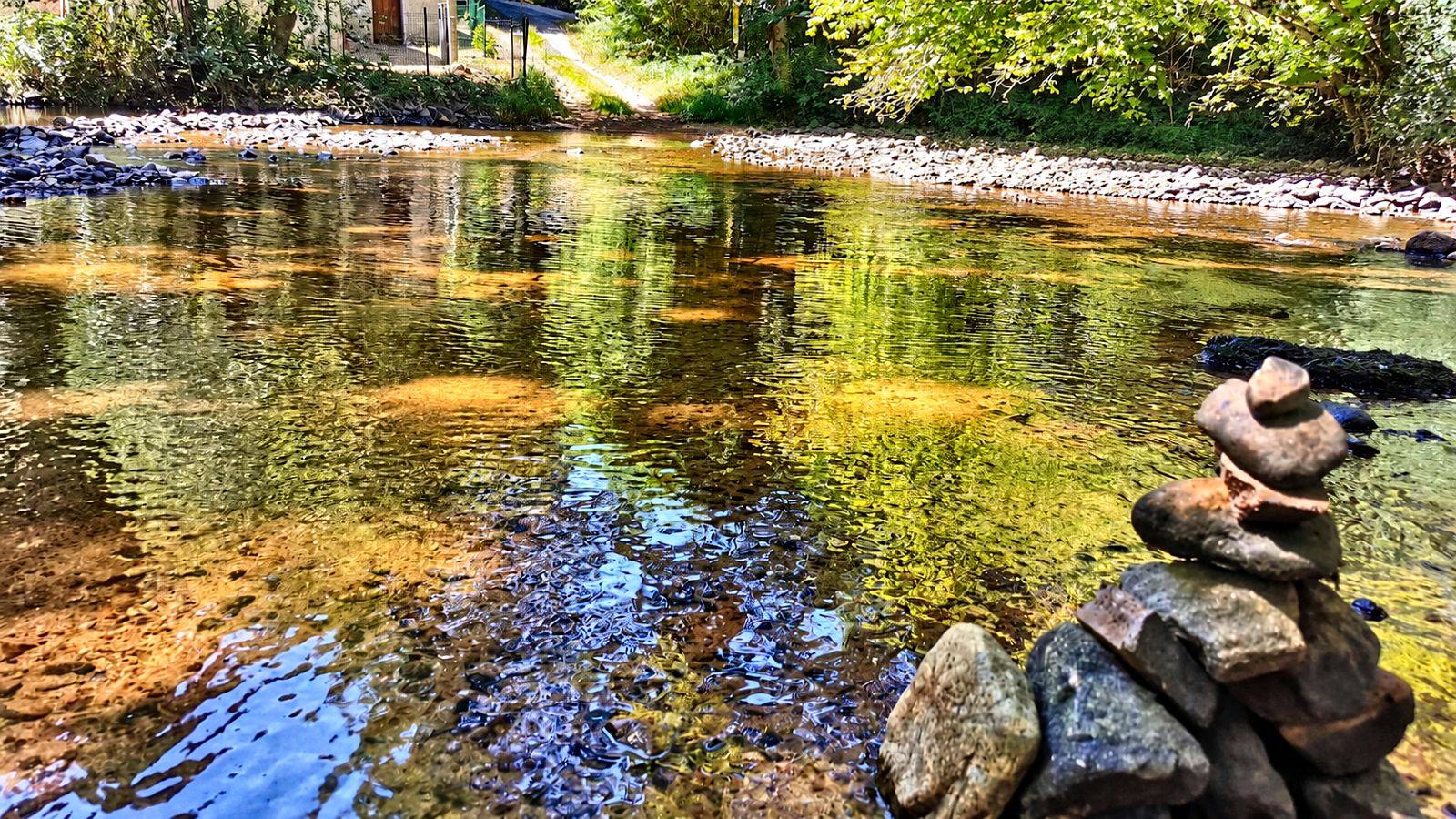 The stream running clear and shallow over golden stones, trees reflected in the surface, with a small stack of balanced stones on the near bank.